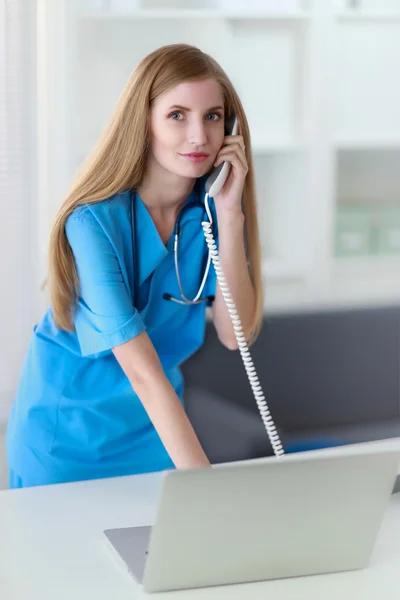 Young woman doctor in white coat at computer using phone — Stock Photo, Image