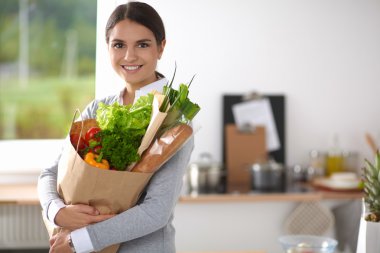 Sebzeli alışveriş poşeti taşıyan genç bir kadın mutfakta duruyor..