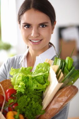 Sebzeli alışveriş poşeti taşıyan genç bir kadın mutfakta duruyor..