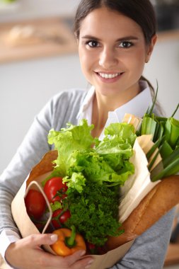 Sebzeli alışveriş poşeti taşıyan genç bir kadın mutfakta duruyor..