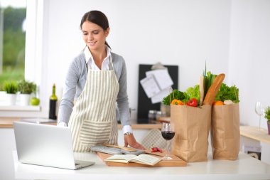 Güzel genç bayan yemek pişiriyor. Mutfakta makbuzu olan dizüstü bilgisayara bakıyor.