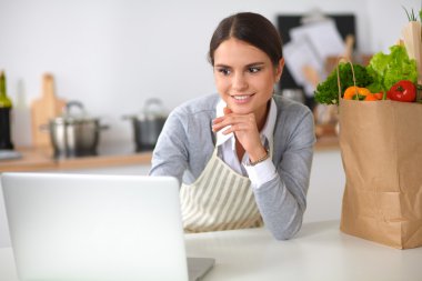 Güzel genç bayan yemek pişiriyor. Mutfakta makbuzu olan dizüstü bilgisayara bakıyor.