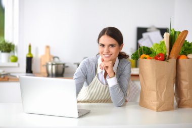 Güzel genç bayan yemek pişiriyor. Mutfakta makbuzu olan dizüstü bilgisayara bakıyor.