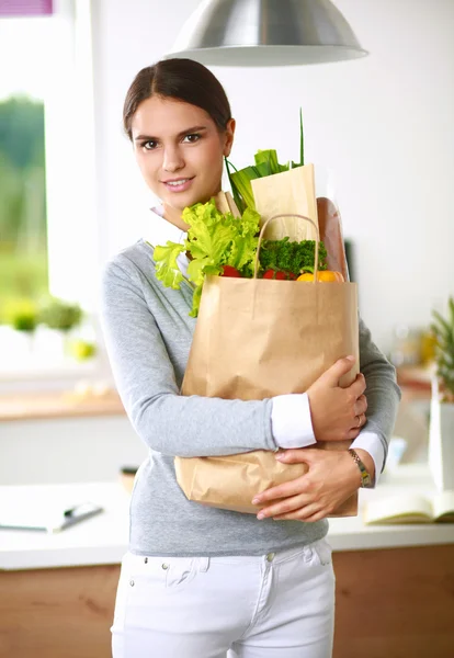 Mujer joven sosteniendo bolsa de la compra de comestibles con verduras de pie en la cocina. —  Fotos de Stock