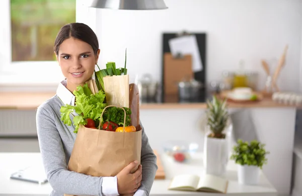 Mujer joven sosteniendo bolsa de la compra de comestibles con verduras de pie en la cocina. —  Fotos de Stock