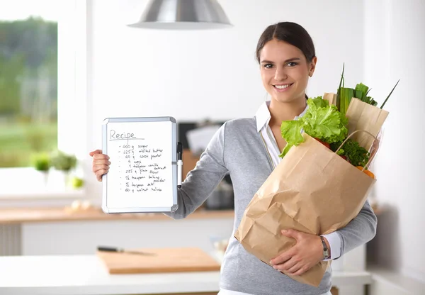 Mujer joven sosteniendo bolsa de la compra de comestibles con verduras de pie en la cocina. —  Fotos de Stock