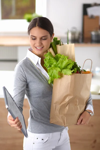 Mujer joven sosteniendo bolsa de la compra de comestibles con verduras de pie en la cocina. —  Fotos de Stock