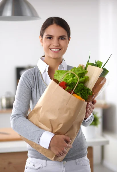 Mujer joven sosteniendo bolsa de la compra de comestibles con verduras de pie en la cocina. —  Fotos de Stock