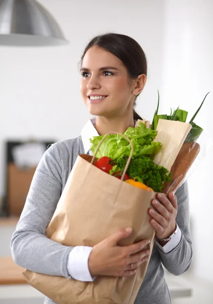 Mujer joven sosteniendo bolsa de la compra de comestibles con verduras de pie en la cocina. —  Fotos de Stock