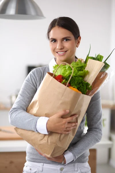 Mujer joven sosteniendo bolsa de la compra de comestibles con verduras de pie en la cocina. —  Fotos de Stock