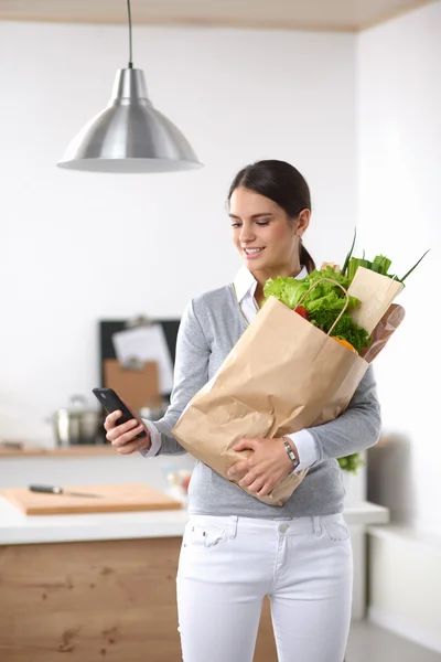 Mujer sonriente con teléfono móvil sosteniendo bolsa de compras en la cocina —  Fotos de Stock