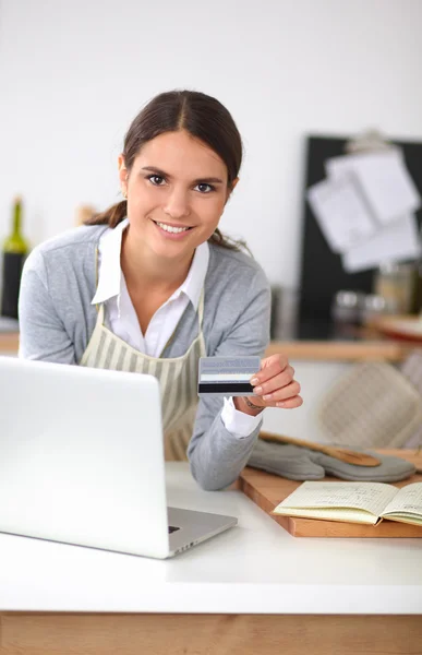 Mujer sonriente compras en línea utilizando la computadora y la tarjeta de crédito en la cocina —  Fotos de Stock