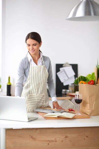 Mooie jonge vrouw koken op zoek naar laptop scherm met ontvangst in de keuken — Stockfoto