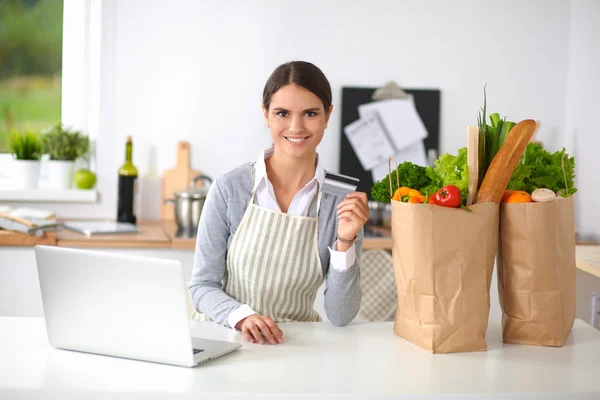 Улыбающаяся женщина совершала интернет-покупки с помощью компьютера и кредитной карты на кухне — стоковое фото