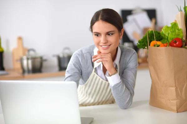 Beautiful young woman cooking looking at laptop screen with receipt in the kitchen — Stock Photo, Image