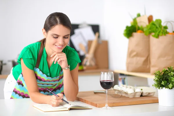 Happy beautiful woman standing in her kitchen writing on a notebook at home — Stock Photo, Image