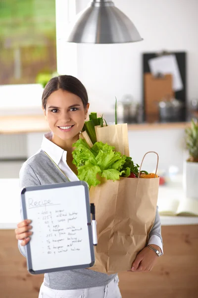 Fiatal nő gazdaság élelmiszerbolt bevásárló táska zöldségekkel Állva a konyhában. — Stock Fotó