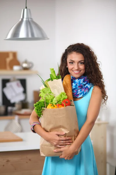 Mujer joven sosteniendo bolsa de la compra de comestibles con verduras de pie en la cocina. — Foto de Stock