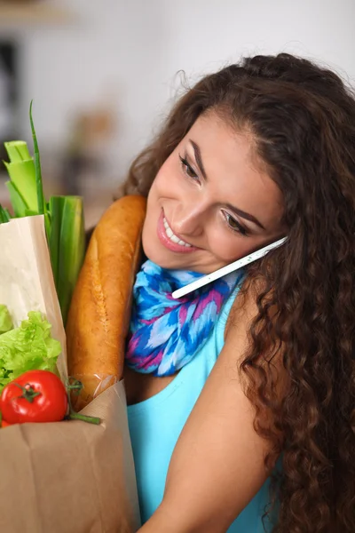 Smiling woman with mobile phone holding shopping bag in kitchen — Stock Photo, Image