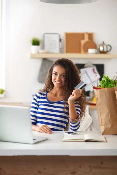 Mujer sonriente compras en línea utilizando la computadora y la tarjeta de crédito en la cocina — Foto de Stock