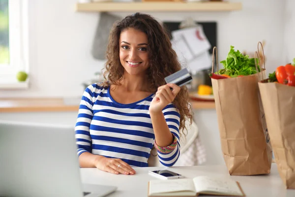 Mujer sonriente compras en línea utilizando la computadora y la tarjeta de crédito en la cocina — Foto de Stock