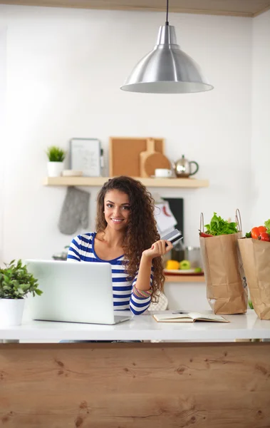 Mujer sonriente compras en línea utilizando la computadora y la tarjeta de crédito en la cocina —  Fotos de Stock