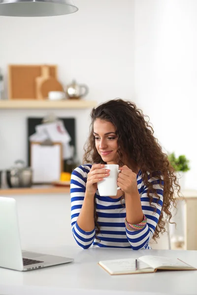 Smiling young woman with coffee cup and laptop in the kitchen at home