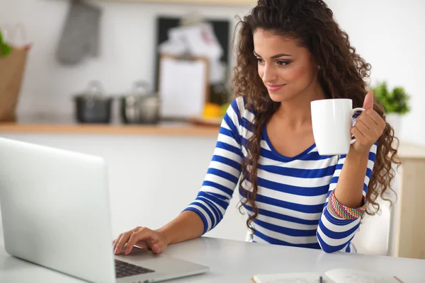 Lachende jonge vrouw met koffiekopje en laptop in de keuken thuis — Stockfoto