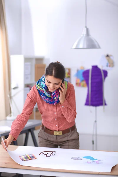 Ofis masasında çalışan genç, çekici bayan moda tasarımcısı, cep telefonuyla konuşurken çizim yapıyor. — Stok fotoğraf