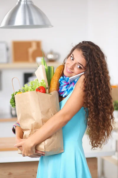 Młoda kobieta trzyma torbę na zakupy z warzywami Stojąc w kuchni. — Zdjęcie stockowe