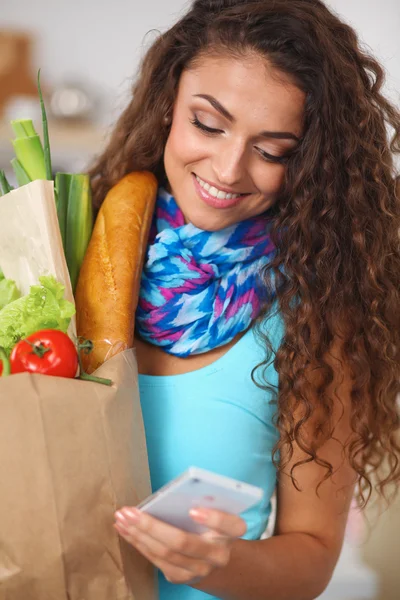 Mujer sonriente con teléfono móvil sosteniendo bolsa de compras en la cocina — Foto de Stock