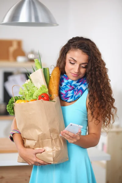 Mujer sonriente con teléfono móvil sosteniendo bolsa de compras en la cocina — Foto de Stock