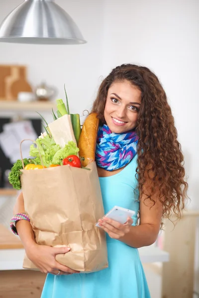 Glimlachende vrouw met mobiele telefoon houden boodschappentas in de keuken — Stockfoto