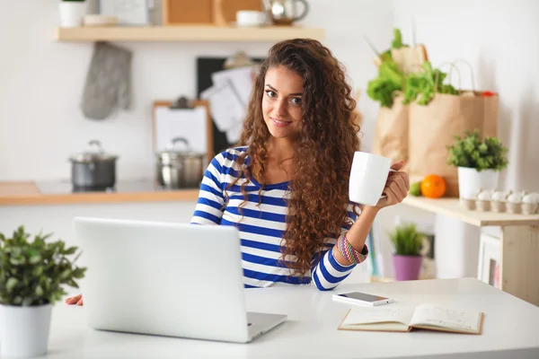 Uśmiechnięta młoda kobieta z filiżanki kawy i laptopa w kuchni w domu — Zdjęcie stockowe