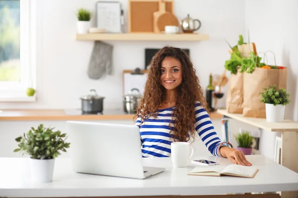 Lachende jonge vrouw met koffiekopje en laptop in de keuken thuis — Stockfoto