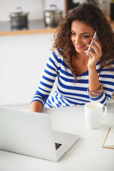 Portrait of a young pretty woman talking the phone and working on laptop in kitchen — Stock Photo, Image