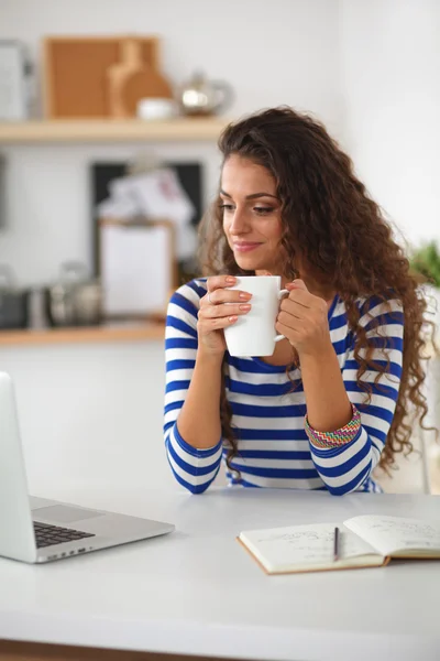 Mujer joven sonriente con taza de café y portátil en la cocina en casa — Foto de Stock