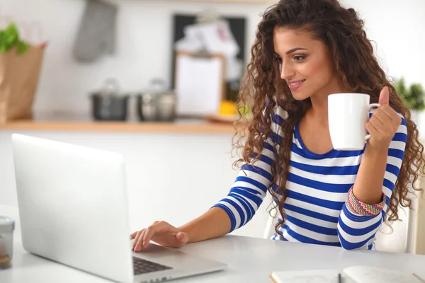 Mujer joven sonriente con taza de café y portátil en la cocina en casa —  Fotos de Stock