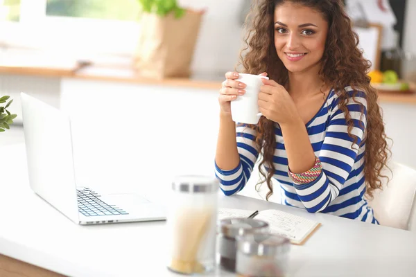 Mujer joven sonriente con taza de café y portátil en la cocina en casa — Foto de Stock