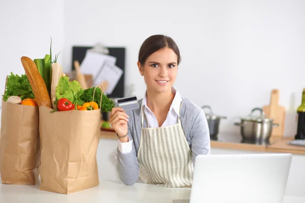 Mulher sorrindo compras on-line usando computador e cartão de crédito na cozinha — Fotografia de Stock