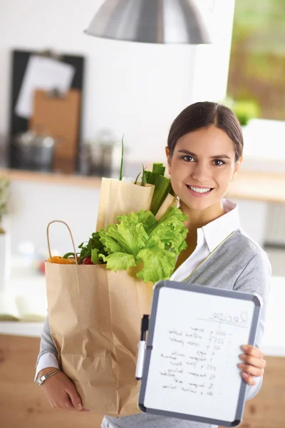 Mujer joven sosteniendo bolsa de la compra de comestibles con verduras de pie en la cocina. — Foto de Stock