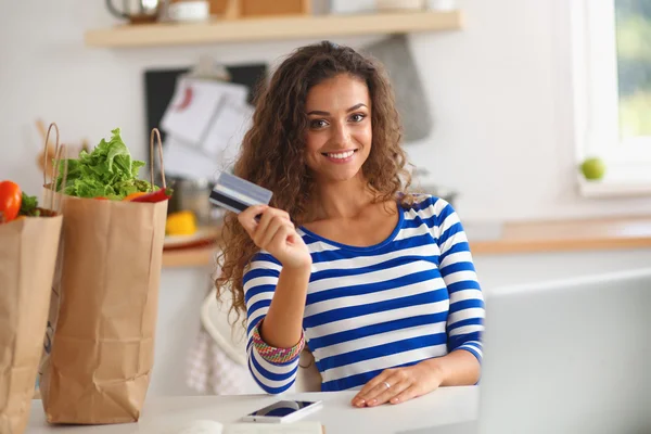 Mujer sonriente compras en línea utilizando la computadora y la tarjeta de crédito en la cocina —  Fotos de Stock