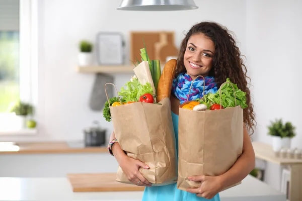 Mujer joven sosteniendo bolsa de la compra de comestibles con verduras de pie en la cocina. — Foto de Stock