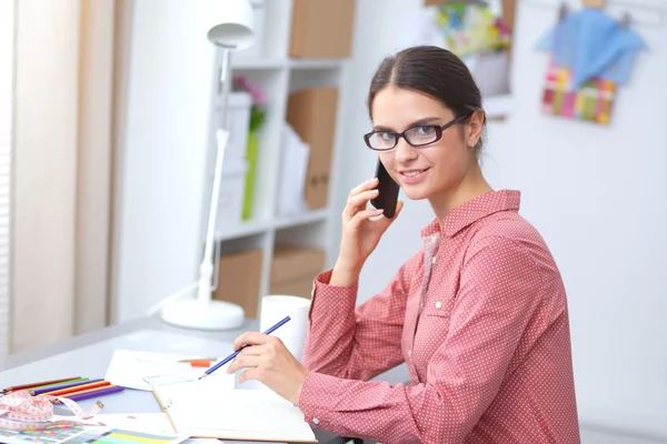 Jonge aantrekkelijke vrouwelijke mode-ontwerper werken op kantoor bureau, tekenen terwijl het praten op mobiele — Stockfoto