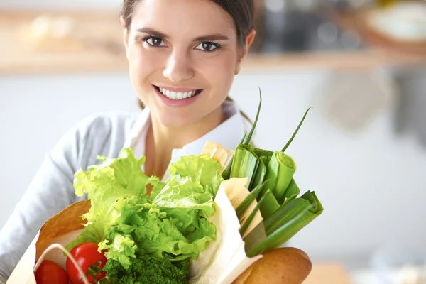 Mujer joven sosteniendo bolsa de la compra de comestibles con verduras de pie en la cocina. —  Fotos de Stock