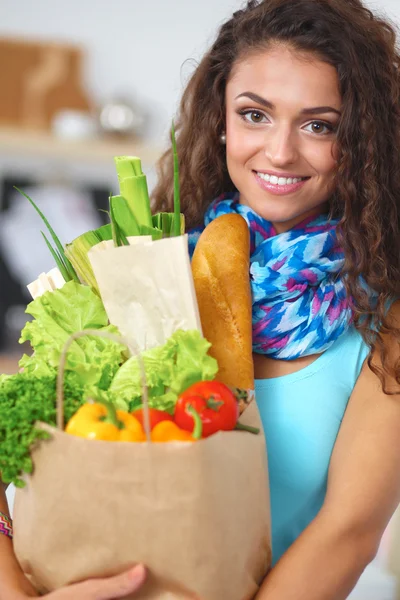 Mujer joven sosteniendo bolsa de la compra de comestibles con verduras de pie en la cocina. — Foto de Stock