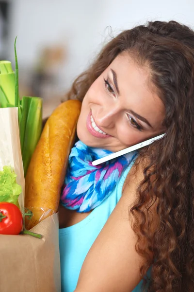 Smiling woman with mobile phone holding shopping bag in kitchen — Stock Photo, Image