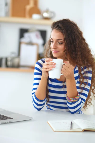 Mujer joven sonriente con taza de café y portátil en la cocina en casa — Foto de Stock