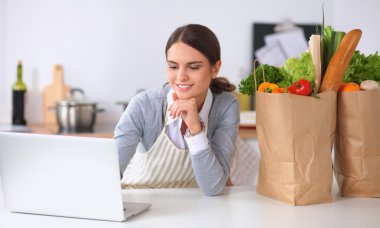 Güzel genç bayan yemek pişiriyor. Mutfakta makbuzu olan dizüstü bilgisayara bakıyor.