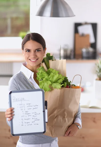 Mujer joven sosteniendo bolsa de la compra de comestibles con verduras de pie en la cocina. —  Fotos de Stock